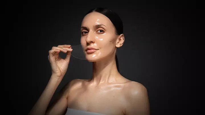 A perfect young woman with sleek dark hair taking the hydrogel mask of her face on dark background A perfect young woman with sleek dark hair taking the hydrogel mask of her face on dark background
